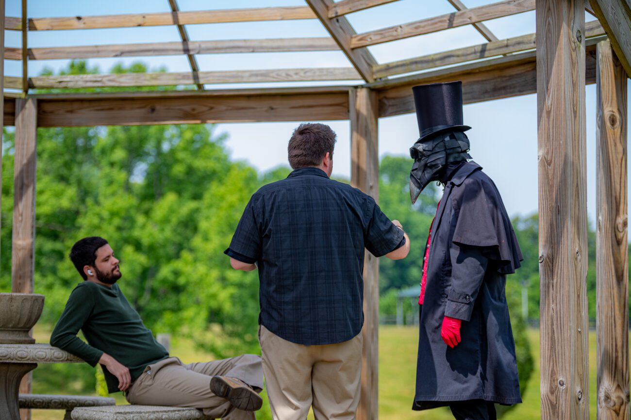 The director discusses a scene with an actor dressed in a plague doctor mask and dark costume. The conversation takes place in a serene outdoor setting, juxtaposing the peaceful environment with the unsettling character design.