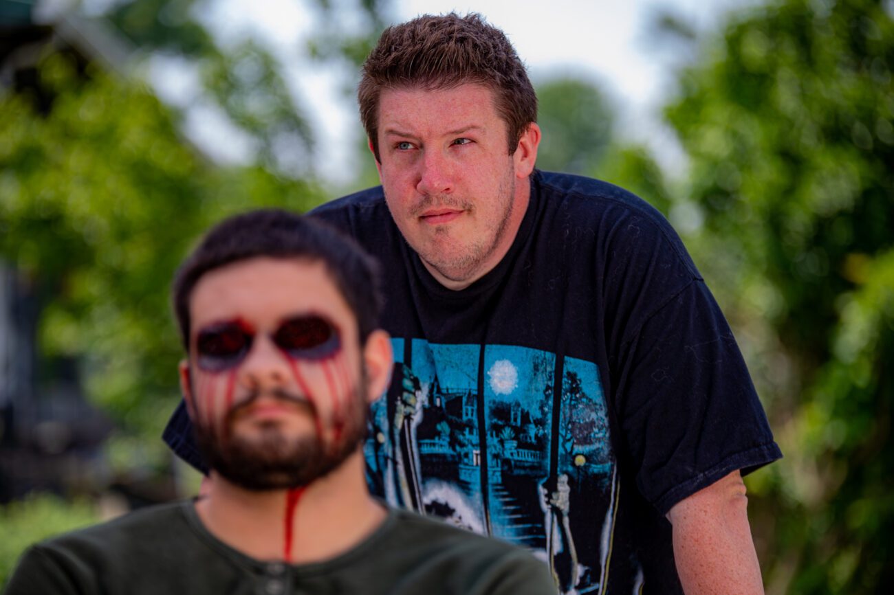 The director stands behind an actor with prosthetic eye makeup creating a gruesome, blood-soaked look. The actor sits calmly in the foreground, while the director looks thoughtfully past him, observing the set.