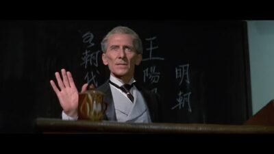 A well dressed older gentlemen with prominent cheek bones gives a lecture with a blackboard behind him.
