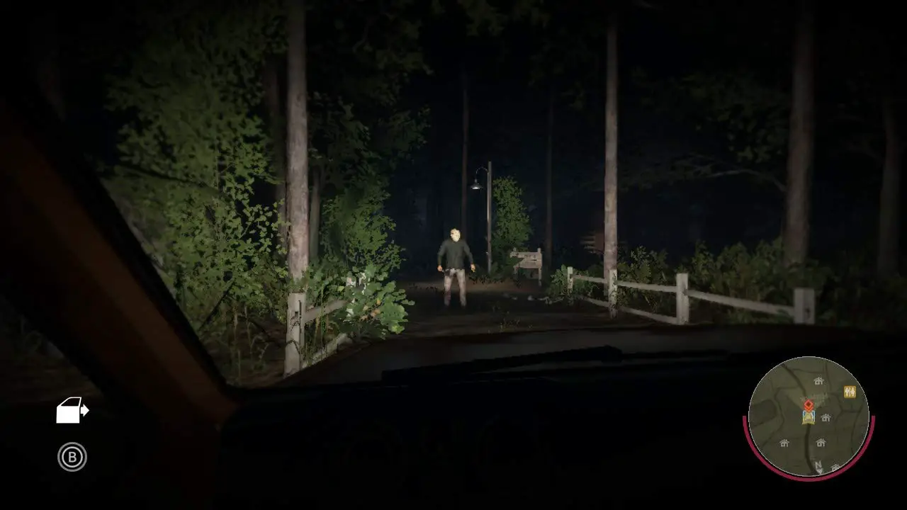 A player’s view from inside a car, headlights casting an glow on Jason Voorhees standing ominously in the middle of a forest road at night, blocking the only way forward.
