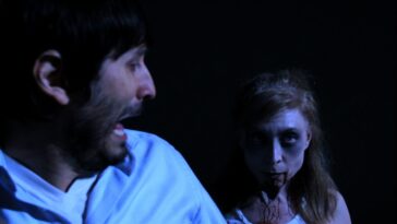A low-light, medium close-up horror movie still from Hell's Kitty. The background is pure black, forcing the focus onto the two actors, illuminated by a pale blue light. On the left, Nicholas Tana (playing Nick, the writer) is shown in profile, mid-scream, wearing a light blue shirt. On the right, horror and adult film icon Nina Hartley (who plays Ms. Rommel in the film) is in full zombie makeup, staring directly at the camera. Her face is pale, eyes are dark and shadowed, and fresh stage blood is smeared around her mouth and neck, giving her a menacing and hungry look. This image captures the jump-scare, nightmare quality of the film's horror elements.